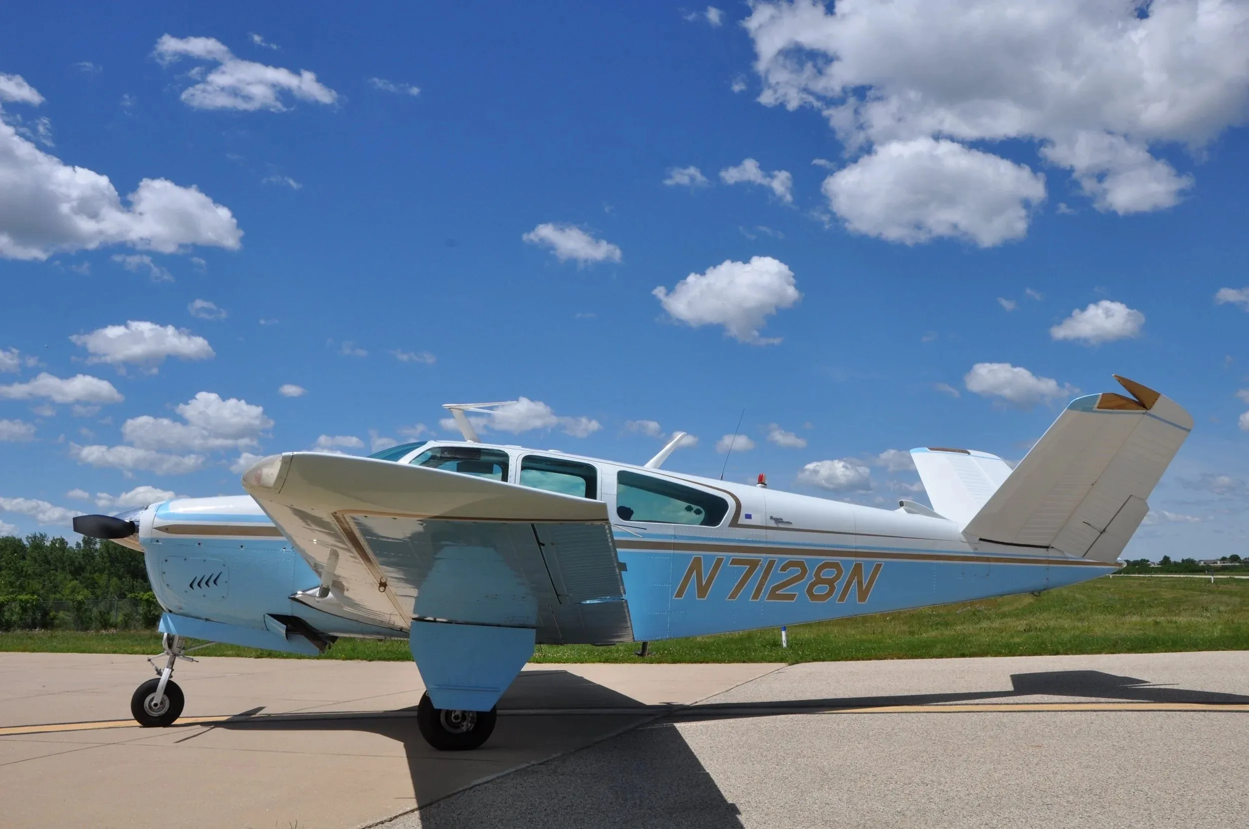 A side view of the light blue and white Beechcraft Bonanza N7128N on an airfield under a cloudy sky.