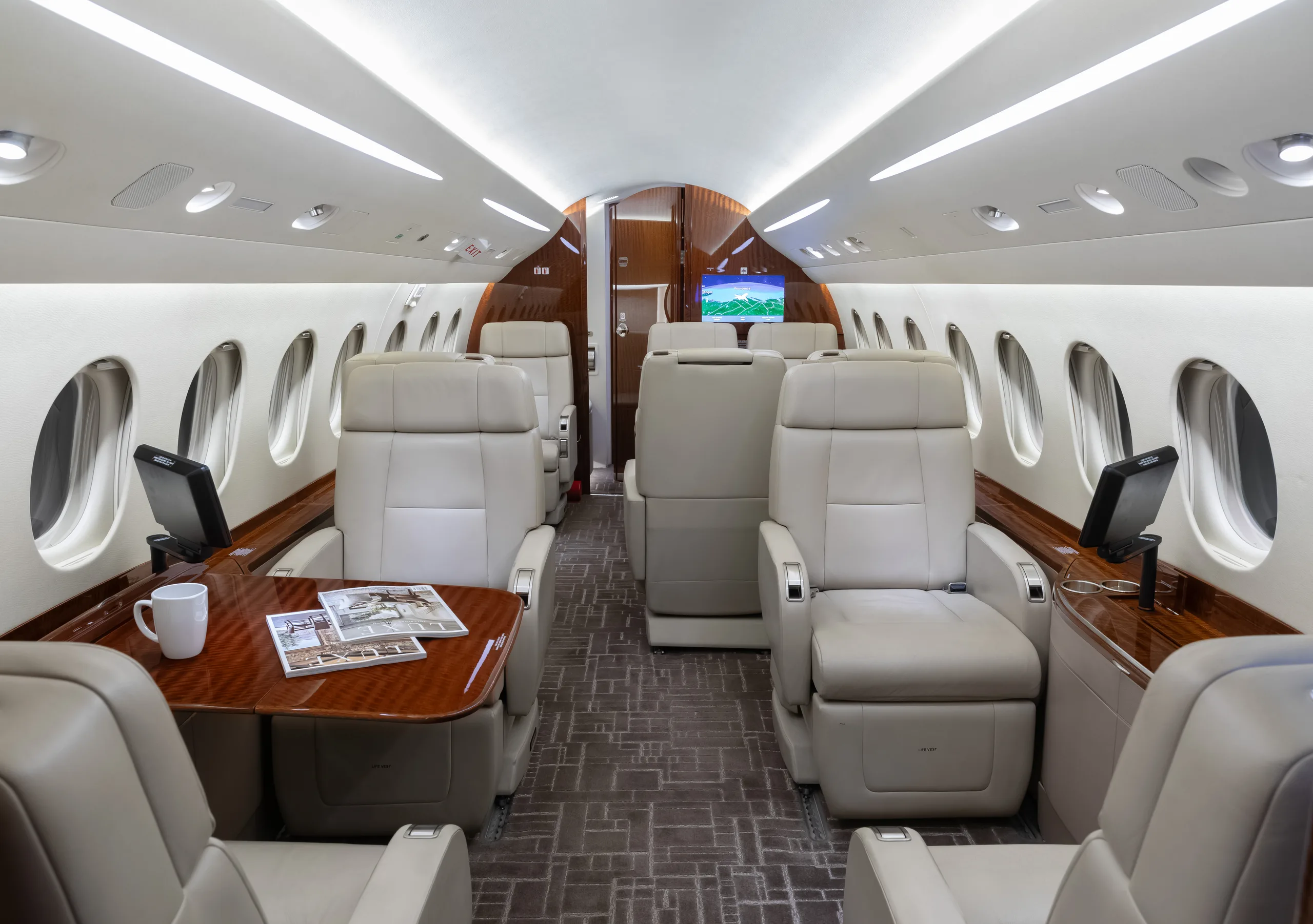 View of a luxurious aircraft cabin with light beige leather seats, a wooden table with magazines and a mug, and a screen at the far end.