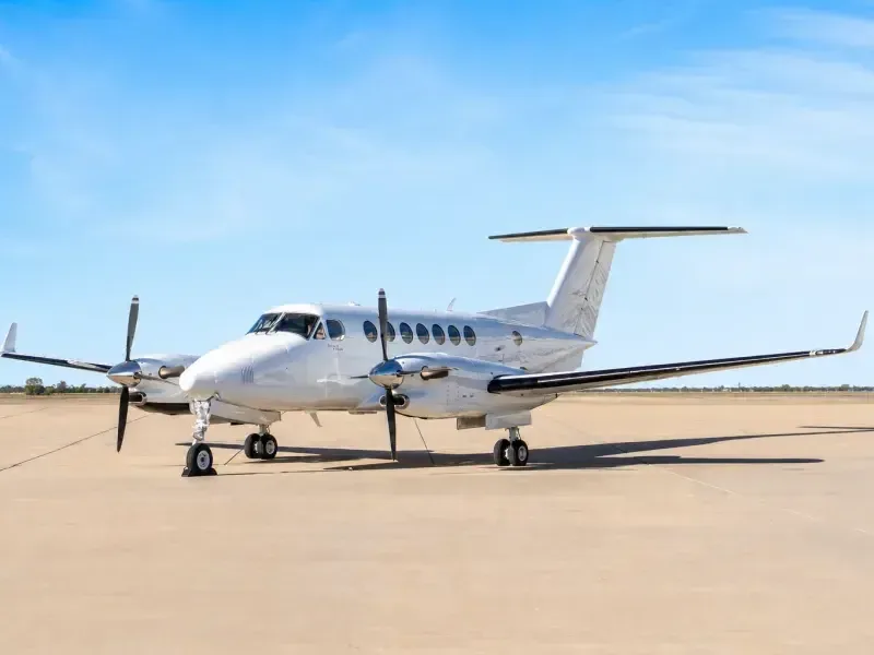 A white Beechcraft King Air twin-turboprop aircraft is parked on an airfield under a clear blue sky.