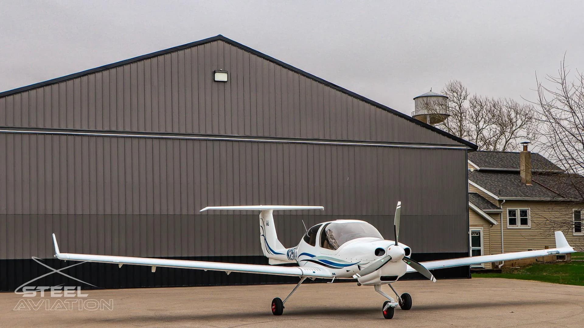 Front 3/4 view of a white Diamond DA40 NG aircraft with blue and grey stripes parked in front of a hangar and a house.