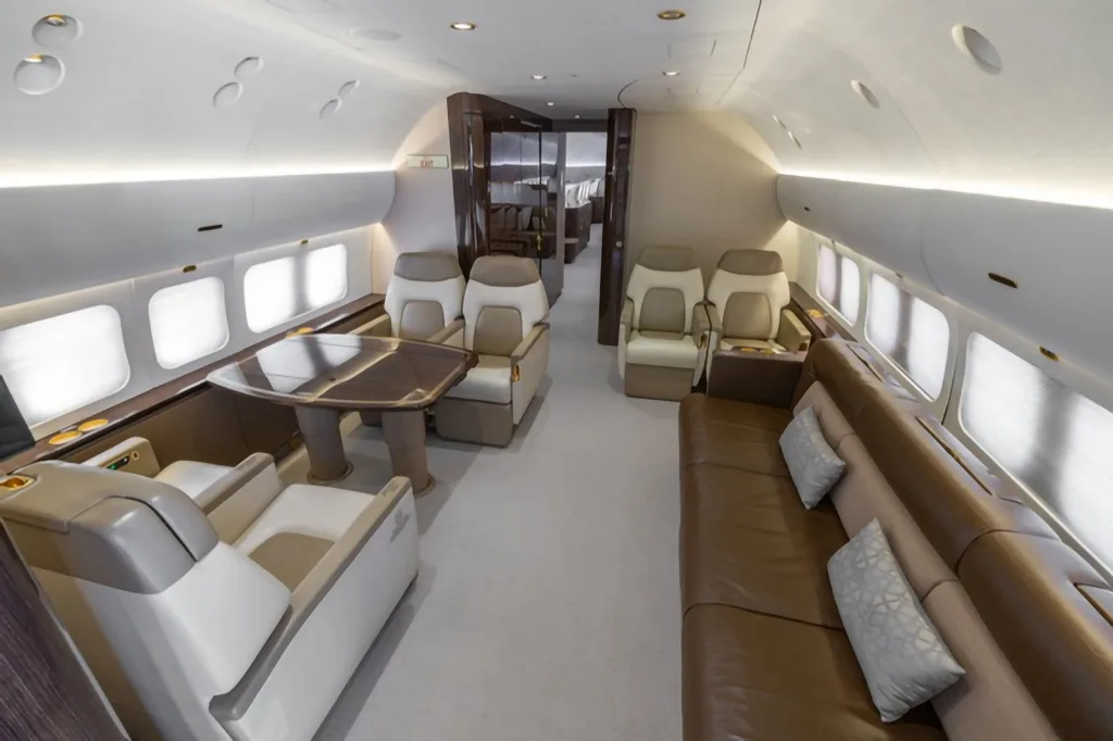 A luxurious aircraft interior featuring beige and brown leather seating, a dining table, and a view towards the rear cabin.