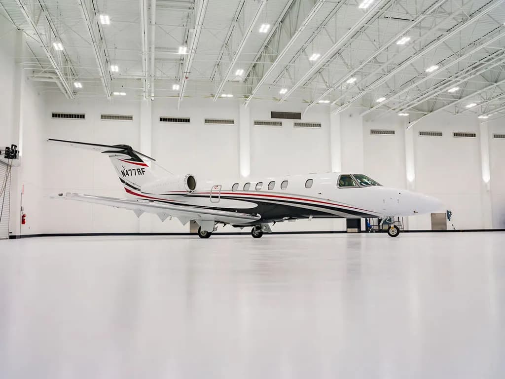 A white Cessna Citation Latitude business jet with black, red, and grey stripes is parked in a brightly lit hangar, viewed from the front-right.