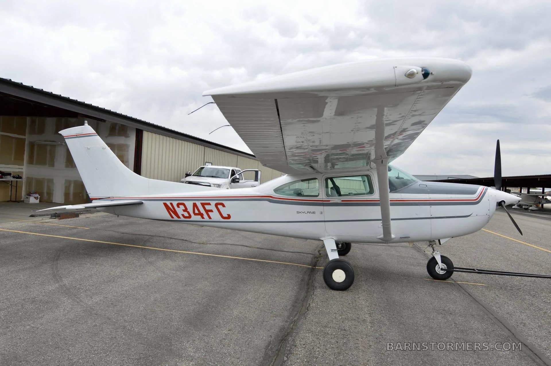A white Cessna 182 Skylane with red and gray stripes, registration N34FC, parked on an asphalt ramp next to a hangar.