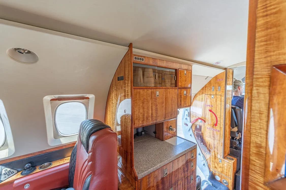 A view of the galley area inside a private jet, featuring rich wood cabinetry and a glimpse into the cockpit.