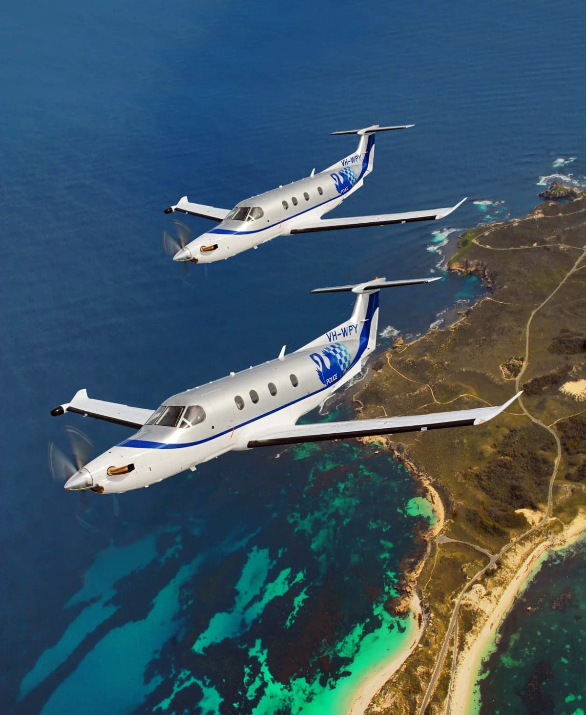 Two Pilatus PC-12 aircraft with police livery fly over a coastline.