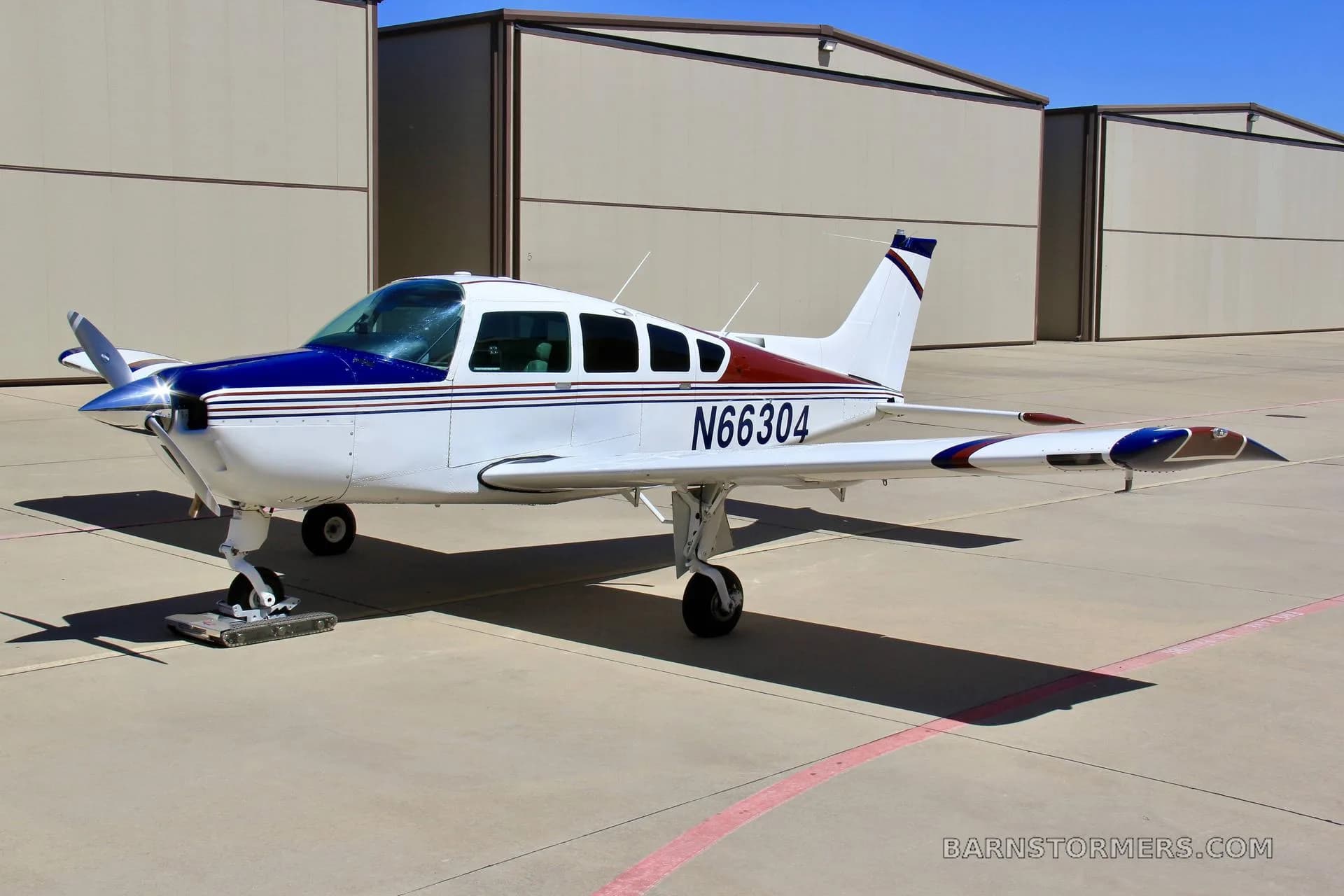 A white single-engine propeller aircraft with blue and red stripes, registration N66304, parked on an airfield in front of hangars.