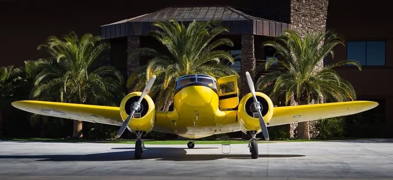 A yellow Beechcraft Model 18 parked in front of a building with palm trees.