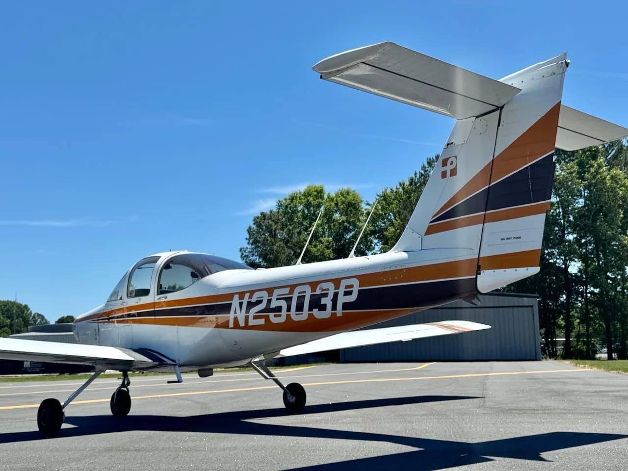 A white Piper PA-38 Tomahawk aircraft with orange and black stripes, seen from the rear-right on an airfield.