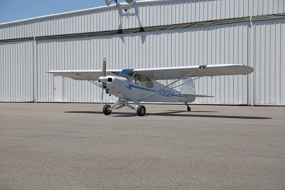 Side view of a white and blue Piper Cub in front of a hangar.