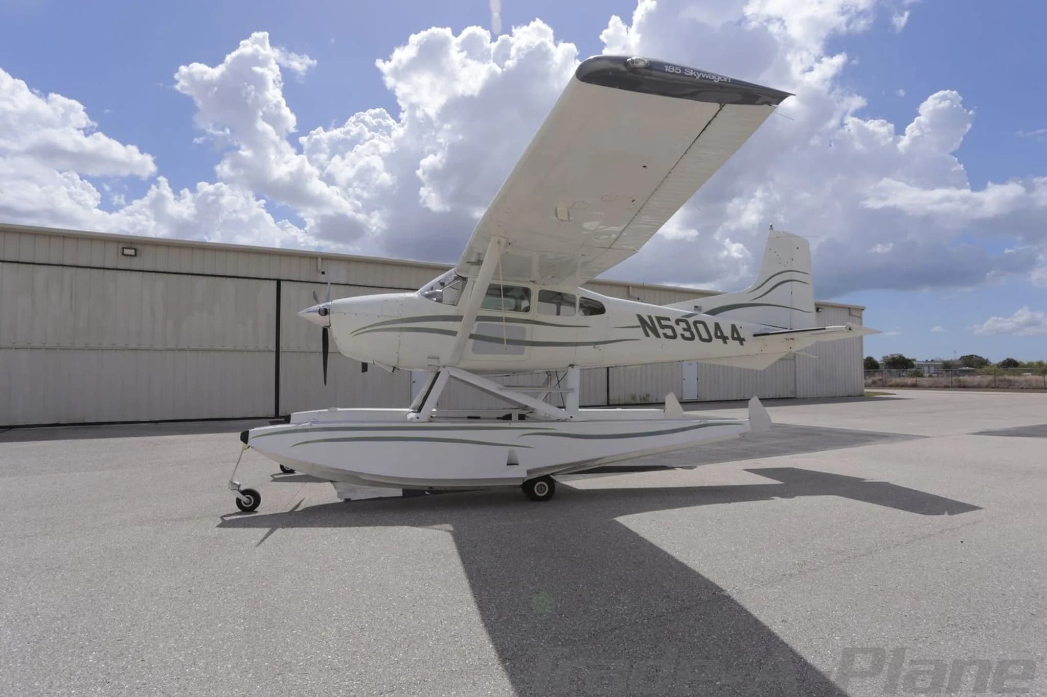A side view of the white Cessna 180 Skywagon seaplane (N53044) with green stripes, parked on an airfield.