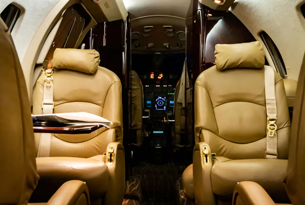 View from the private jet's cabin, showing tan leather seats in the foreground and the illuminated cockpit instrument panel in the distance.