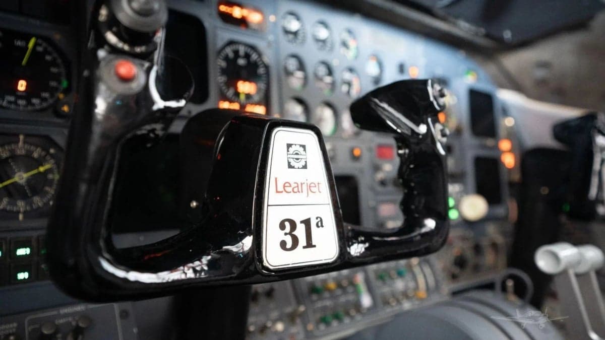 Close-up of a black control yoke with "Learjet 31A" label, in a cockpit with various instruments in the background.