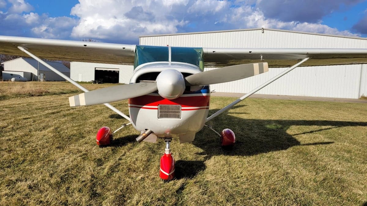 A front view of the Cessna Skylane with red, white, and blue paint scheme on a grassy field in front of a hangar.