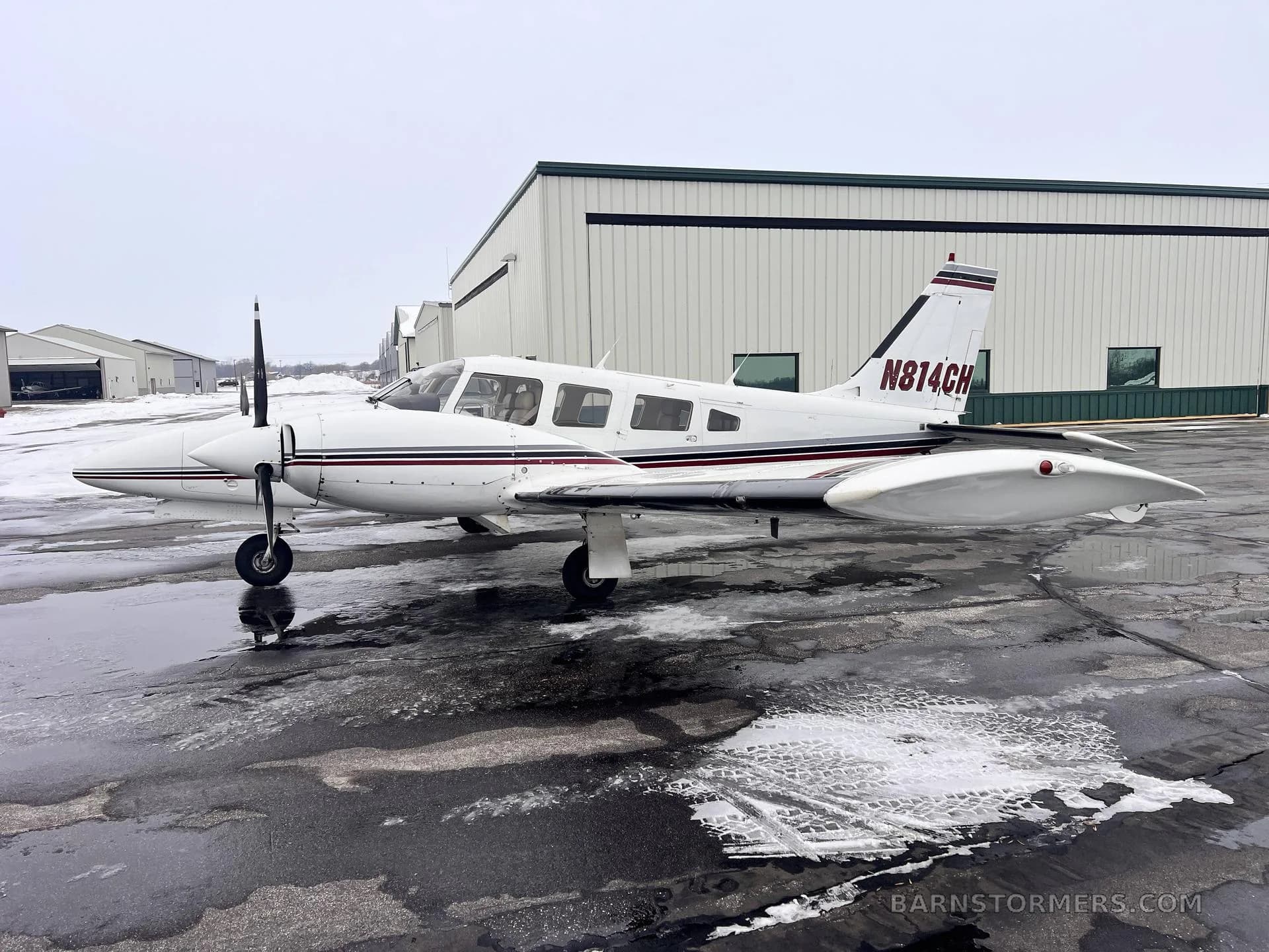 A white twin-engine Piper aircraft with maroon and black stripes, tail number N814CH, parked on a wet tarmac with snow next to a hangar.