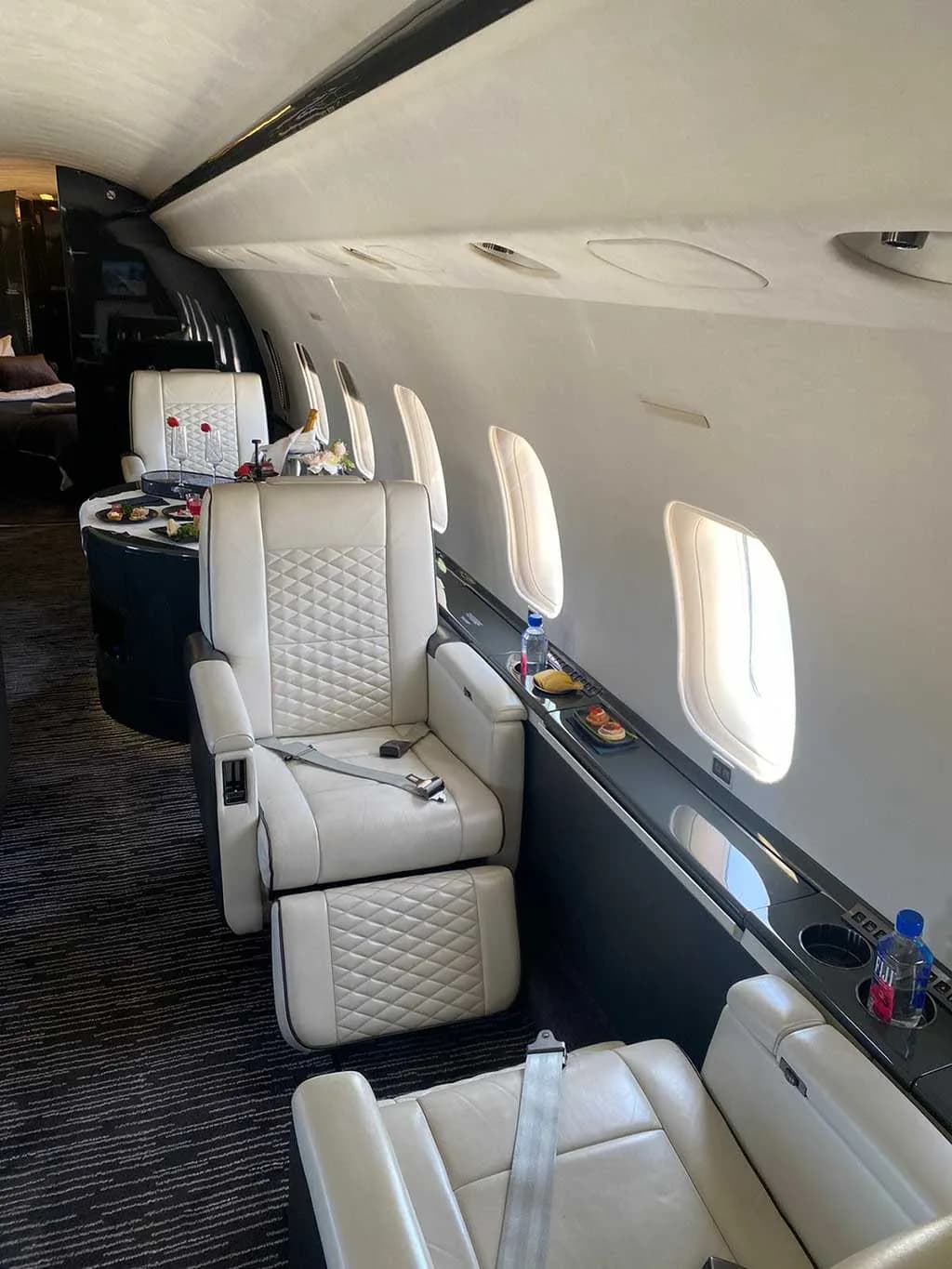 The private jet cabin with white quilted seats, a dark table with snacks and drinks, and large windows along the side.