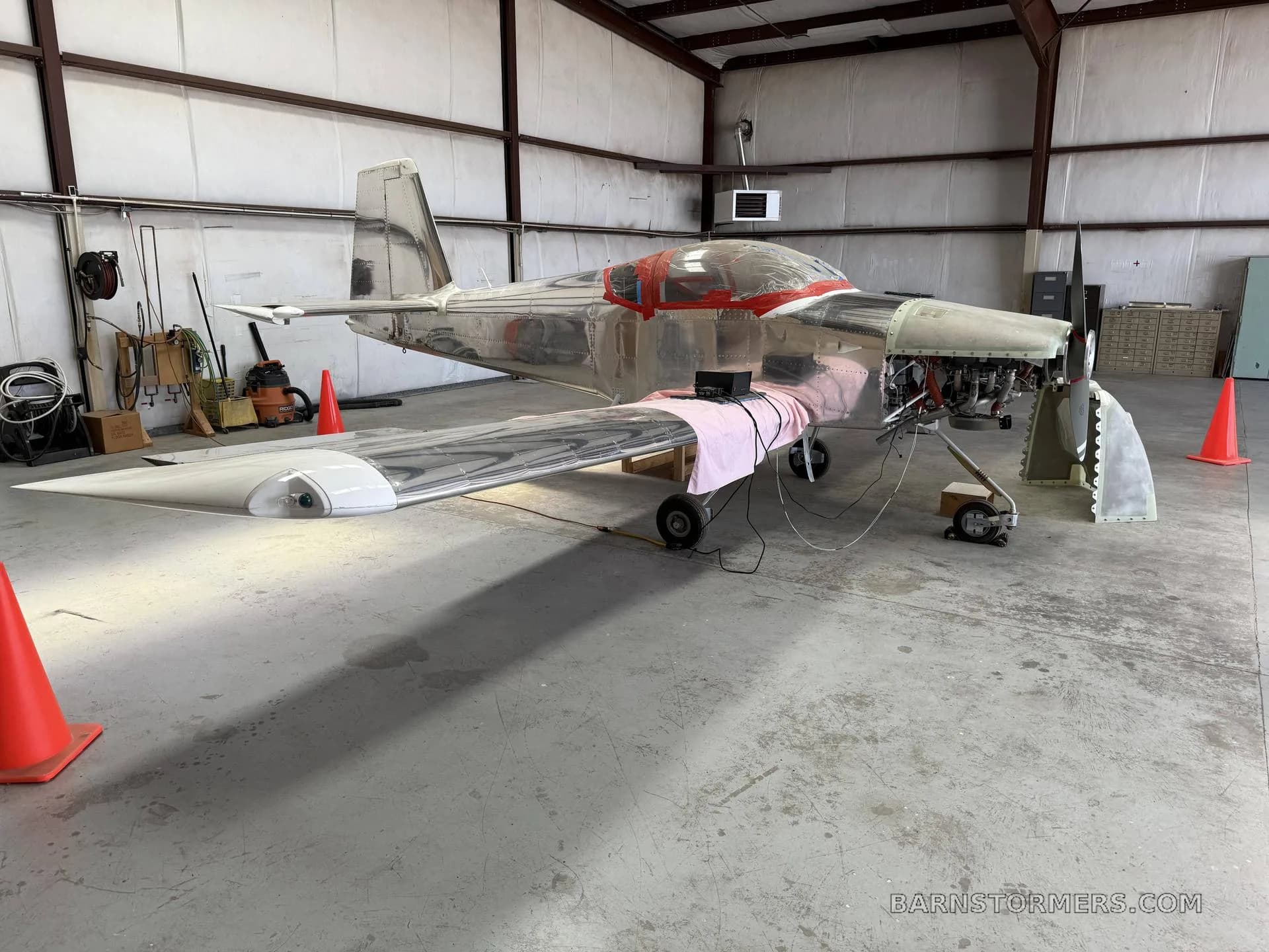 A rear-side view of a partially assembled, unpainted metal aircraft with a red-taped canopy in a hangar.