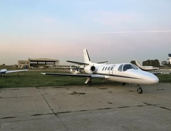 A white business jet with black accents parked on an airfield tarmac at dusk, with a hangar in the background.