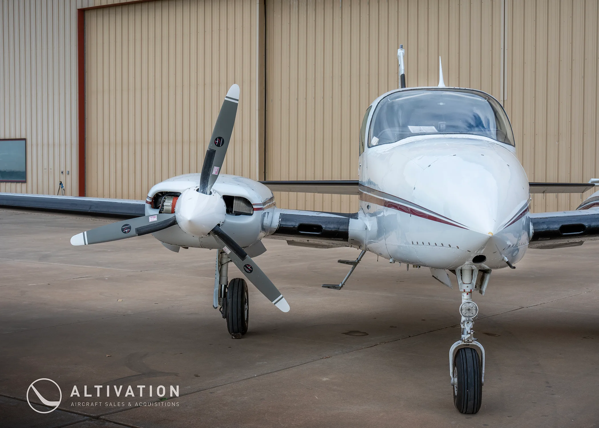 A front view of a white twin-engine propeller aircraft with maroon stripes, tail number N2642Y, parked in front of a hangar.
