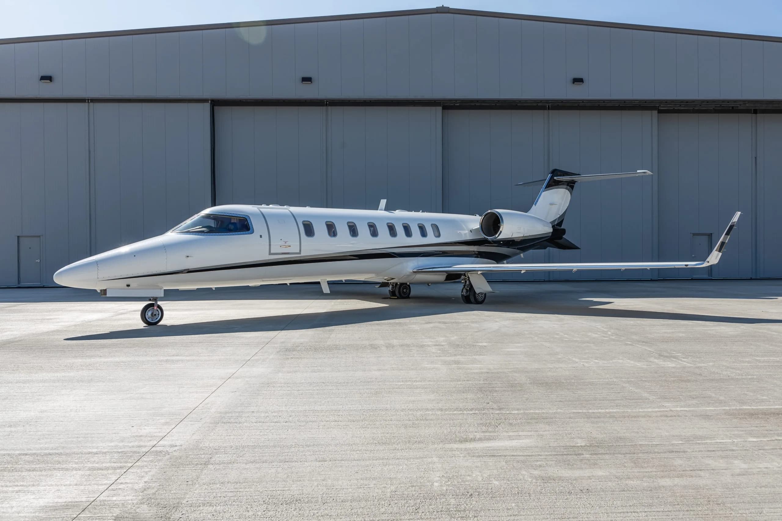 A white business jet with black accents is parked on a tarmac in front of a gray hangar under a clear sky.