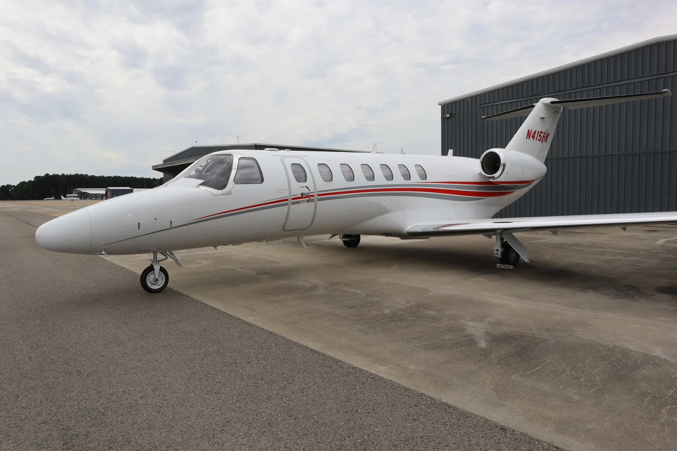 A side view of the white Cessna Citation business jet N415RK with red and grey stripes, parked on an tarmac next to a grey hangar.