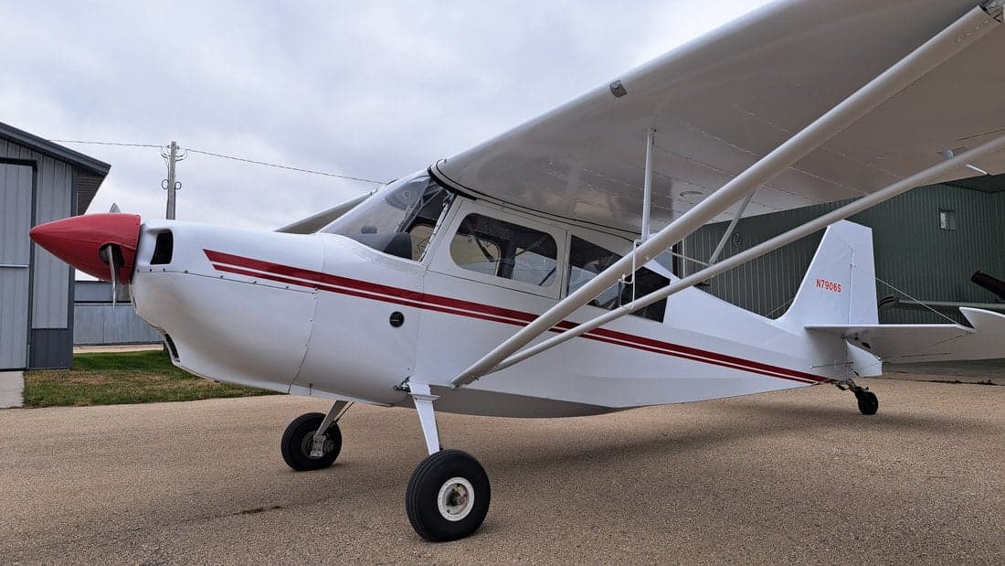 A white single-engine aircraft with red stripes and a red nose is parked on an asphalt surface under a cloudy sky.