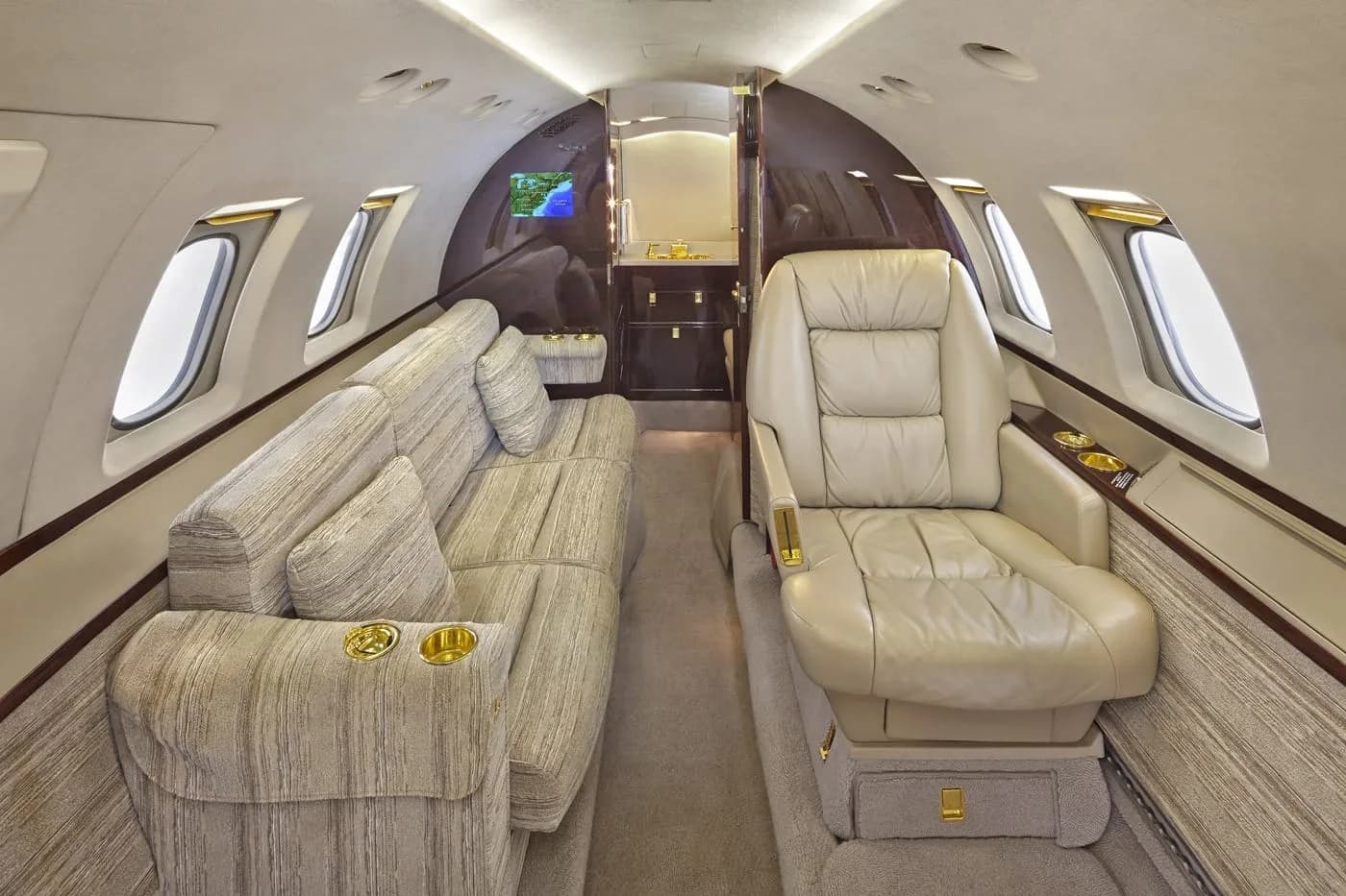The luxurious interior of a private jet cabin, featuring a beige sofa and a single cream leather seat, with a galley/lavatory area in the background.