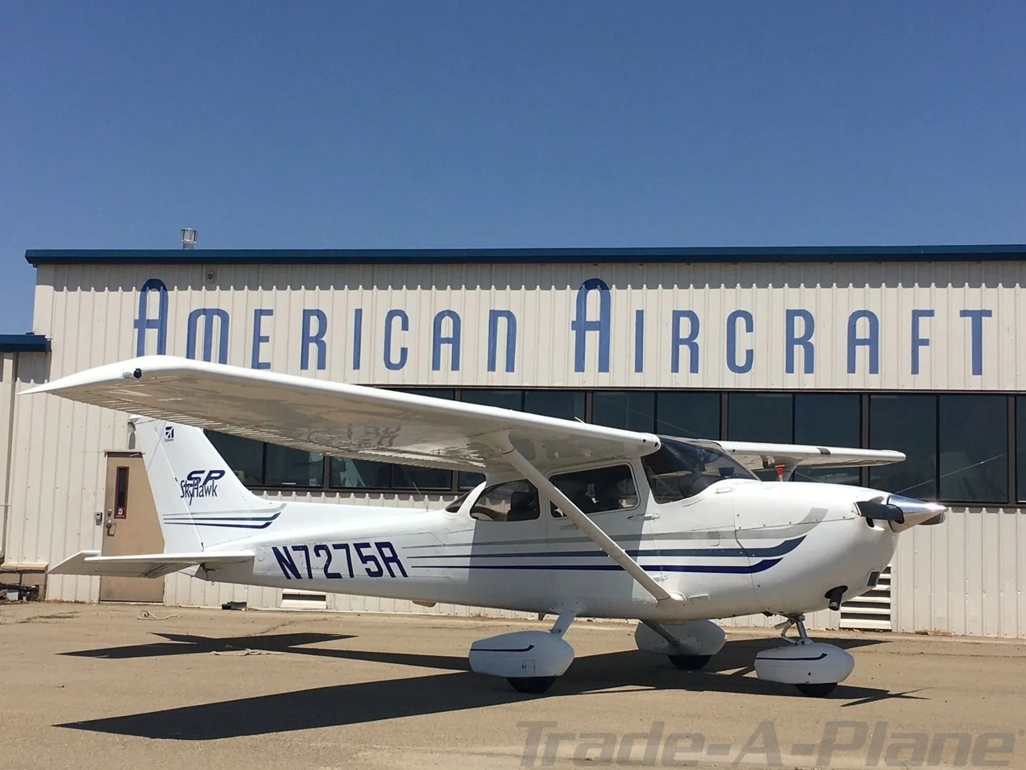 A white Cessna 172SP Skyhawk with blue stripes, registration N7275R, parked in front of a building labeled "AMERICAN AIRCRAFT".
