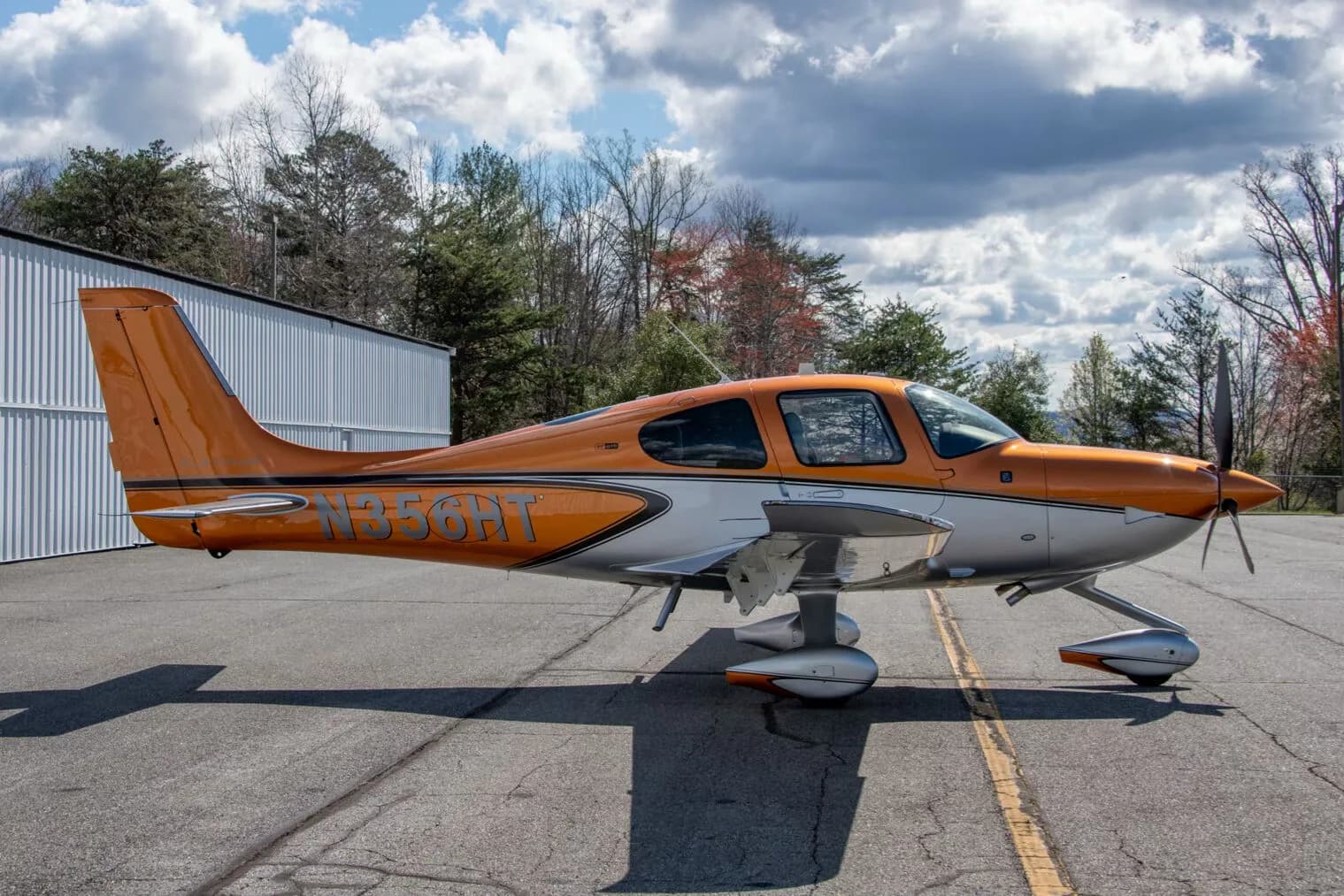 A side view of a metallic orange and silver Cirrus SR22 aircraft (N356HT) parked on an asphalt ramp with trees in the background.