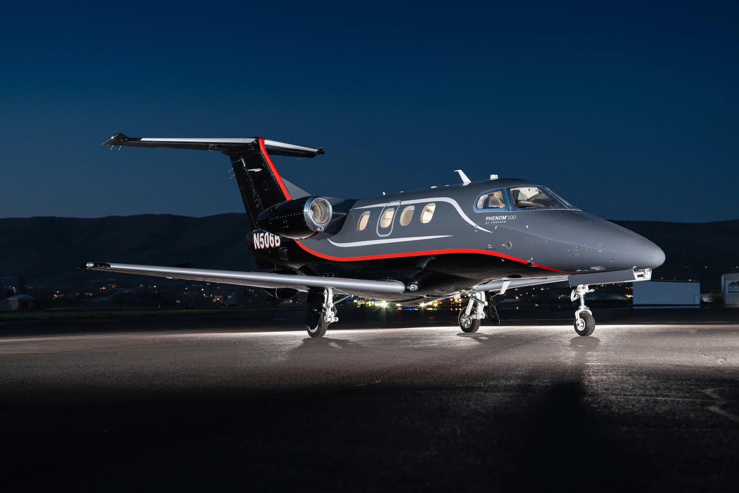 A dark gray and black Embraer Phenom 100 with red and white stripes, registration N506B, parked on a runway at night with mountains in the background.