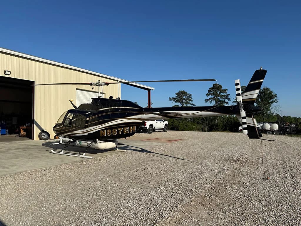 A black and gold helicopter, N887EH, is parked on a gravel lot next to an open hangar.