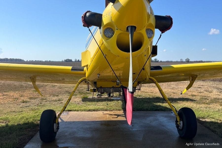 Front view of a yellow agricultural aircraft with a propeller, parked on a concrete pad in a field.