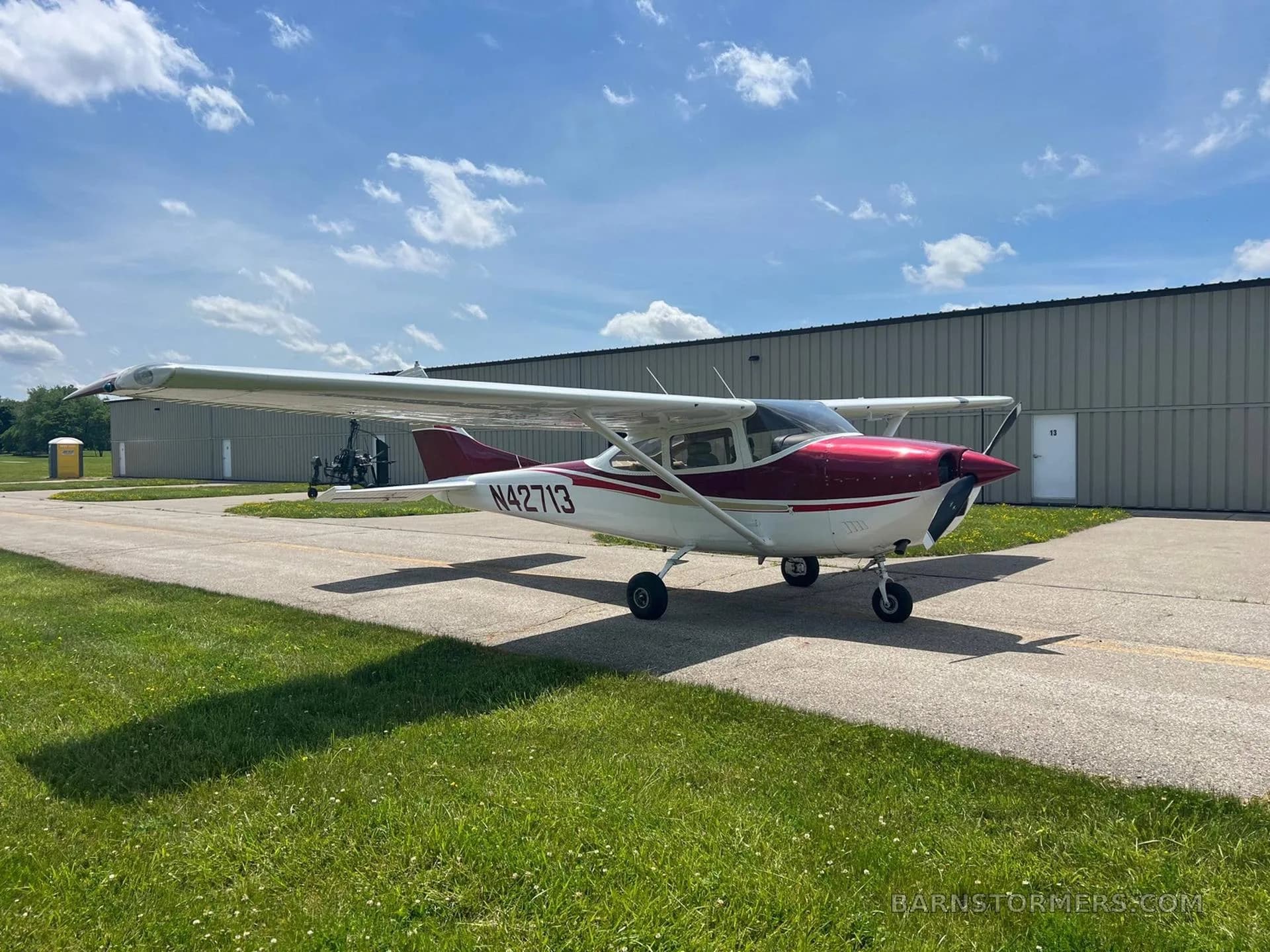 A red and white Cessna N42713 parked on an asphalt taxiway next to a hangar under a partly cloudy sky.