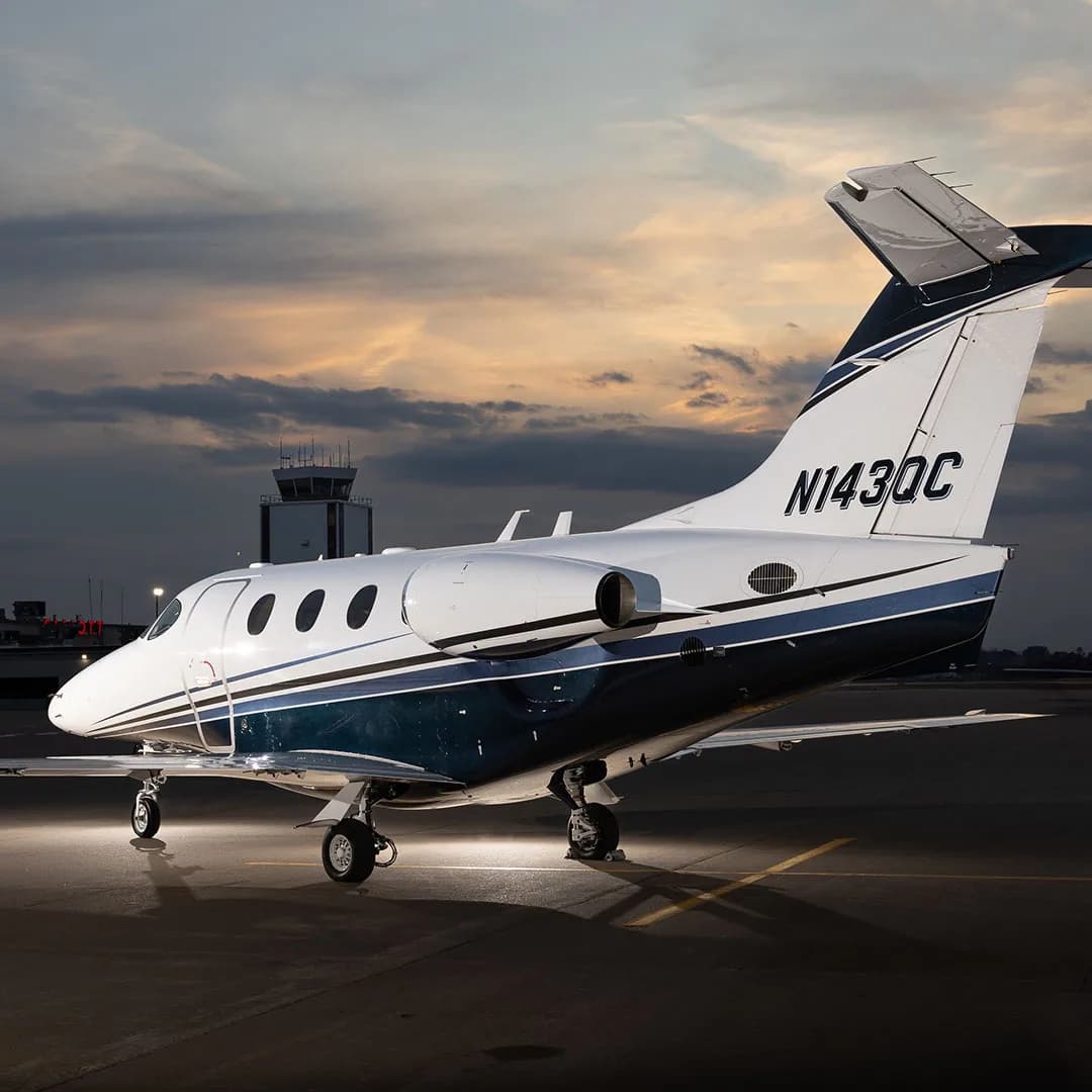 A white and blue business jet, registration N143QC, parked on an airfield at sunset with an air traffic control tower in the background.