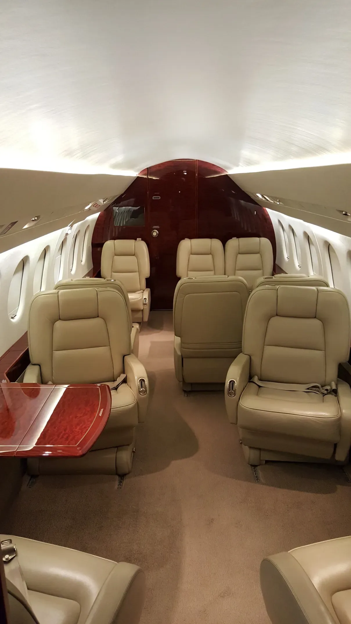The spacious interior cabin of a private jet featuring beige leather seating and a polished wooden table.