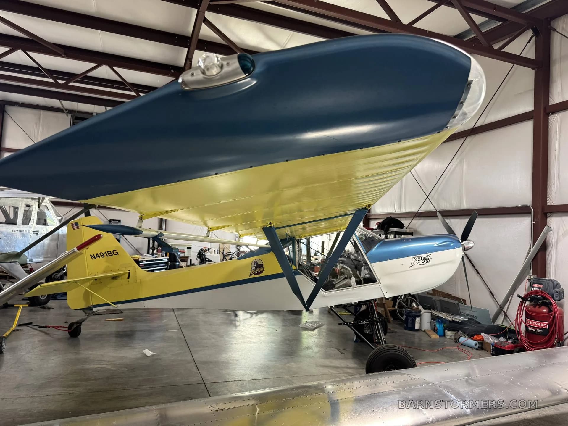 Side view of a yellow and blue Kitfox aircraft with its wing folded, parked in a hangar.