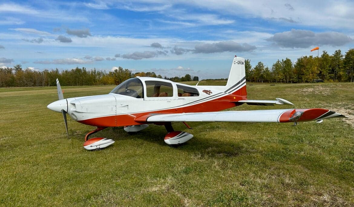A white and orange Grumman American AA-5A Cheetah aircraft is parked on a grassy field under a blue sky.