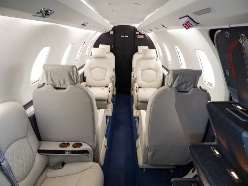 The interior cabin of a business jet, featuring light beige leather seats and a blue carpeted aisle, looking towards the rear.