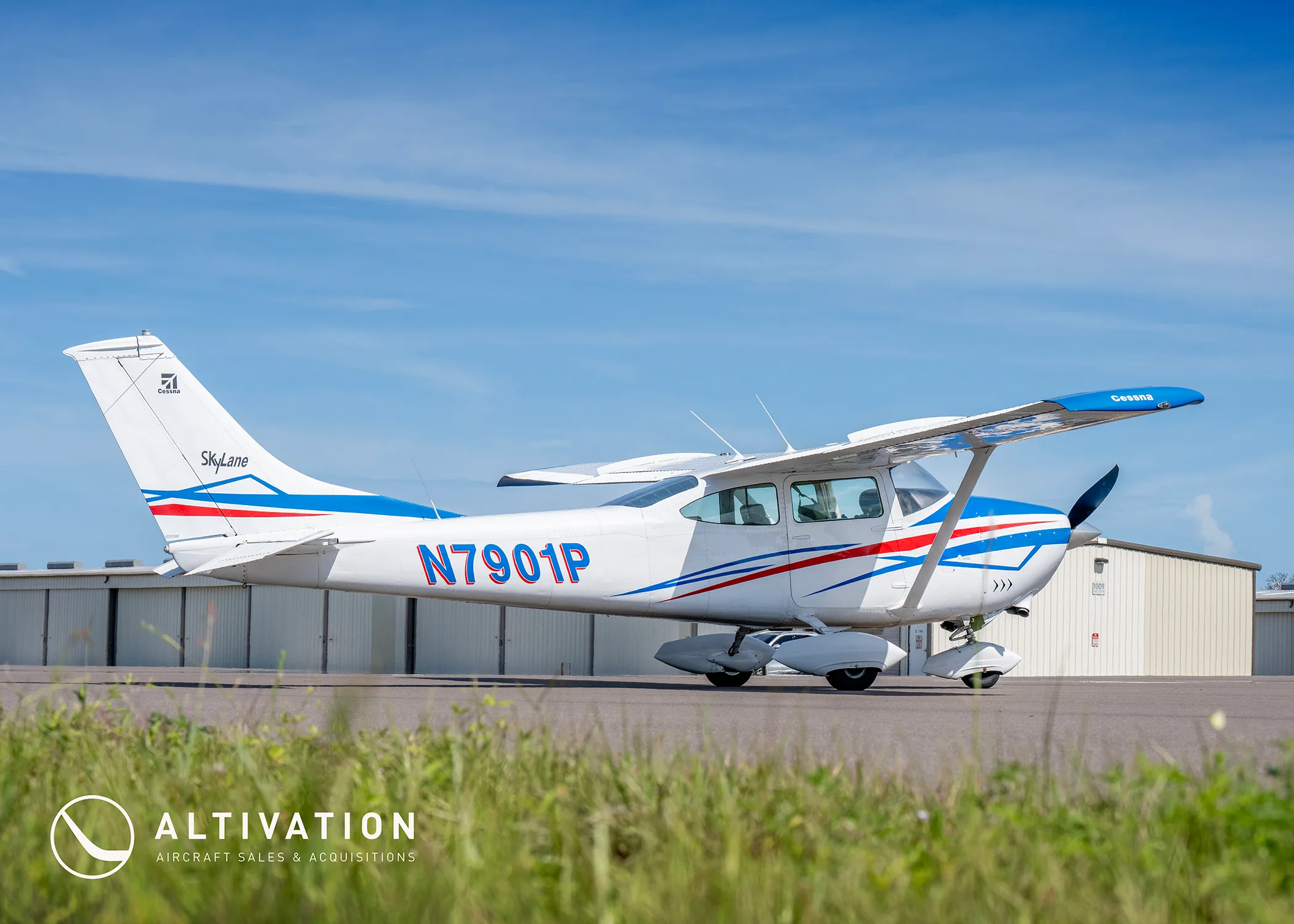 A white Cessna 182 Skylane with blue and red stripes parked on a tarmac with hangars in the background.