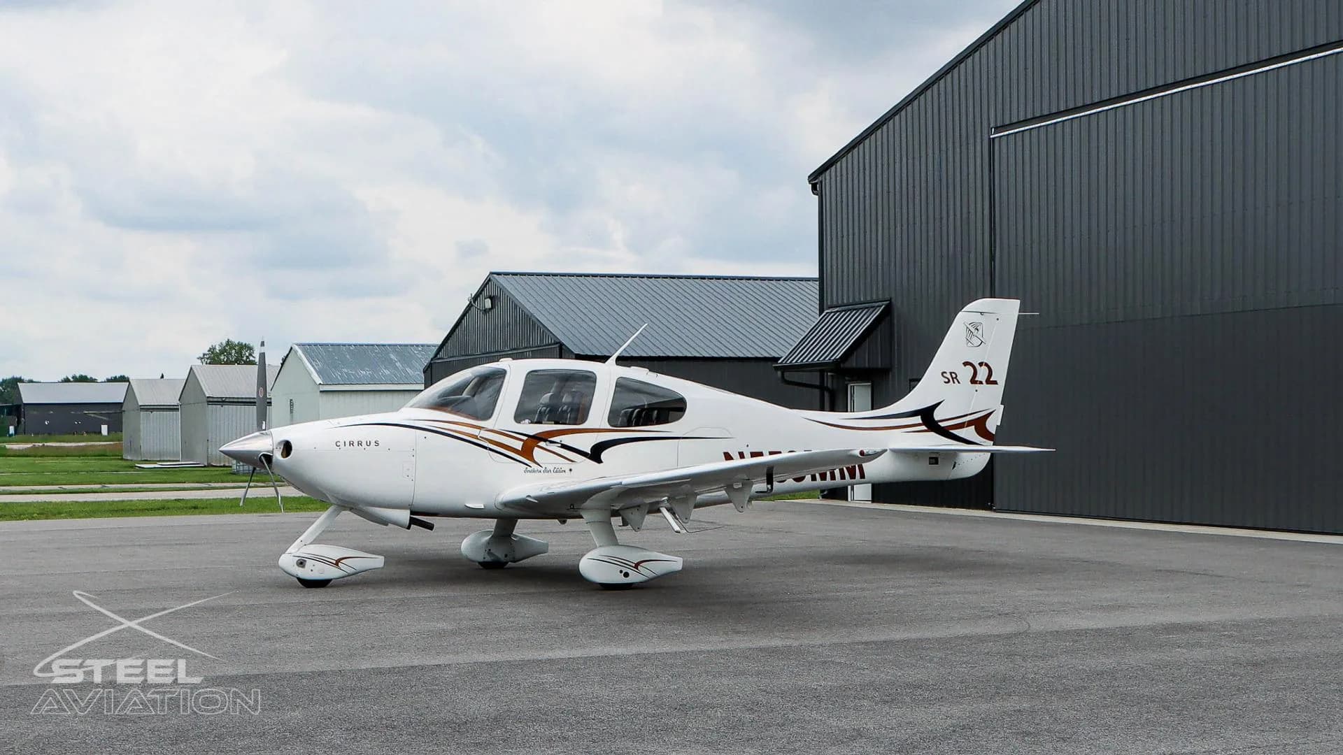 A white Cirrus SR22 aircraft with N559MM registration is parked on an airfield with hangars under a cloudy sky.