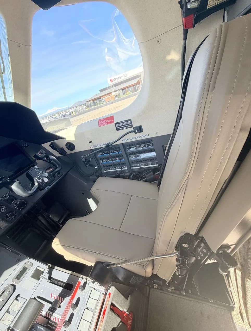 The pilot's seat and side console of a Pilatus PC-12 aircraft, with an airport building visible through the window.