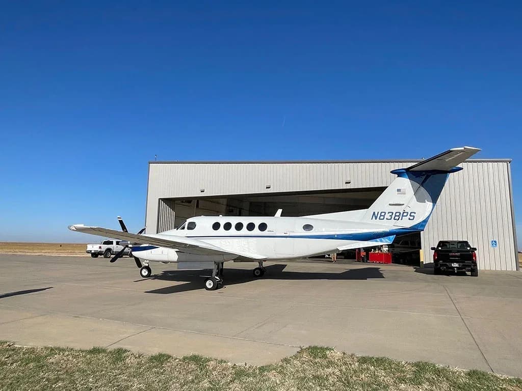 A white and blue twin-engine turboprop aircraft, N838PS, parked on an airfield with a hangar in the background.