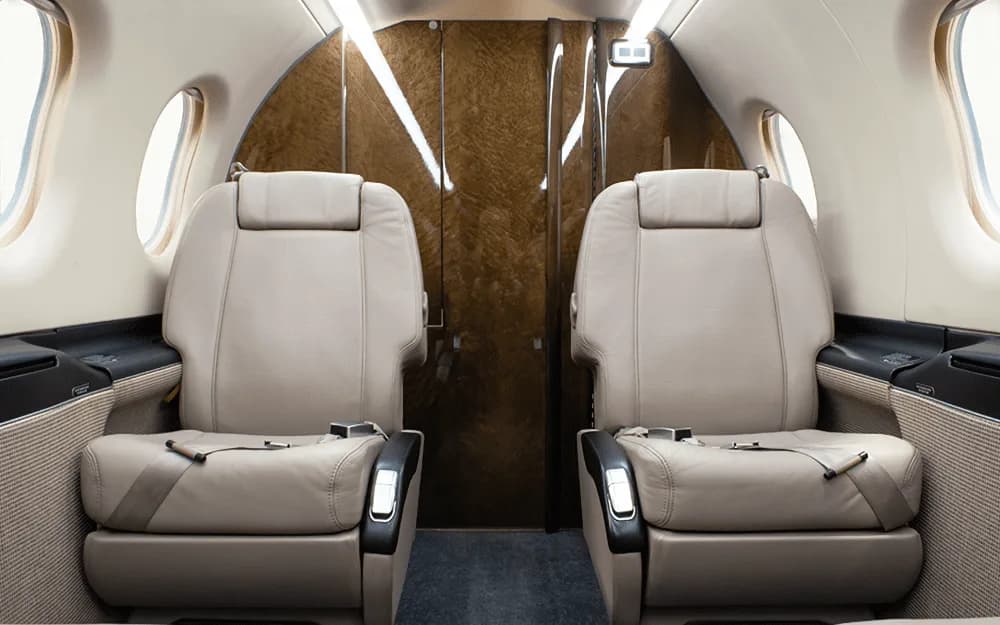 Two beige leather passenger seats in the cabin of a Pilatus PC-12 NG, facing a wood-paneled bulkhead.