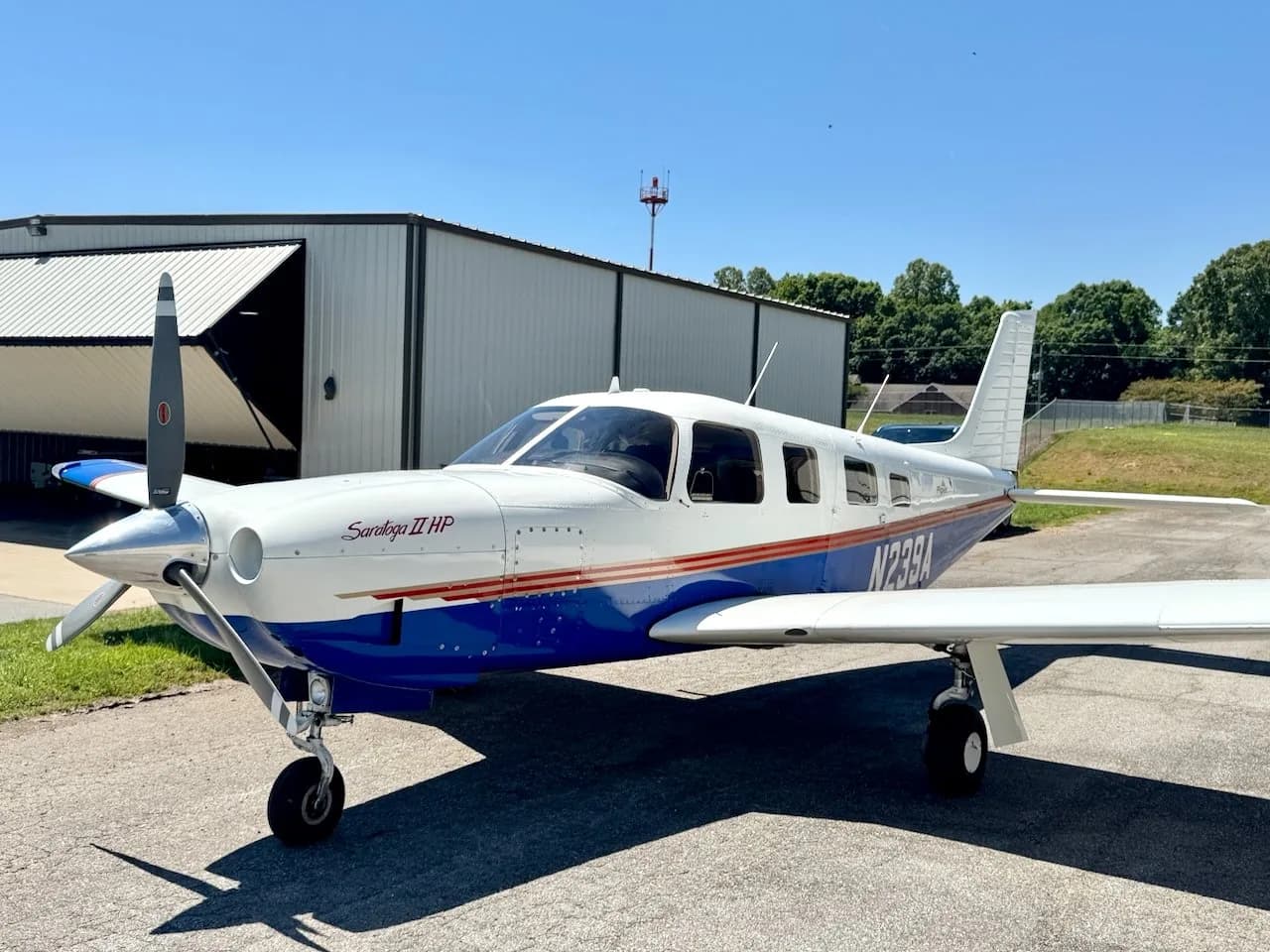 A three-quarter front view of a white and blue Piper Saratoga II HP aircraft, registration N239A, parked on an airfield next to a hangar.