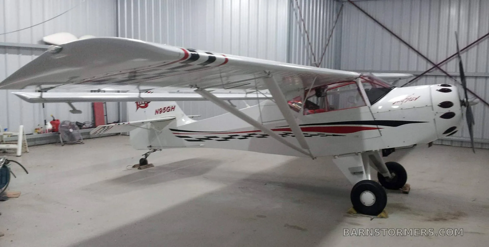 A white SkyStar Kitfox aircraft with red and black checkerboard accents is viewed from the front-right inside a hangar.
