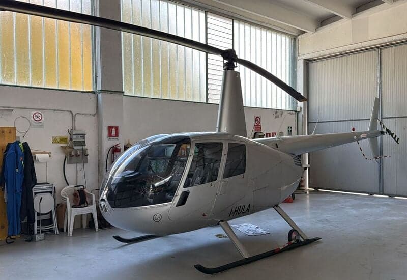 Side view of a silver Robinson R44 helicopter with the registration I-HULA inside a hangar.