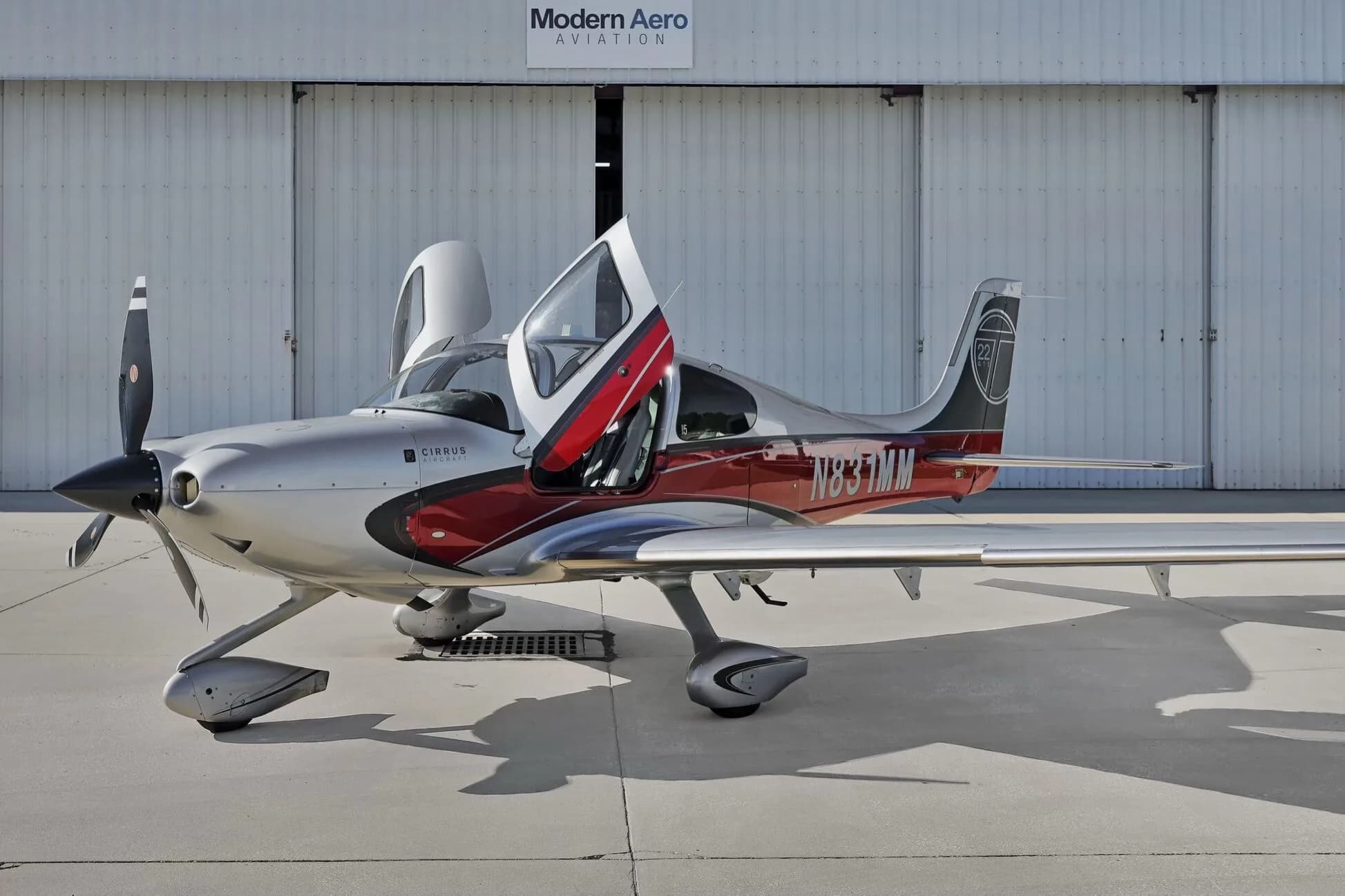 Side view of a Cirrus SR22T aircraft with red and silver paint scheme, registration N831MM, parked on an airfield with doors open.