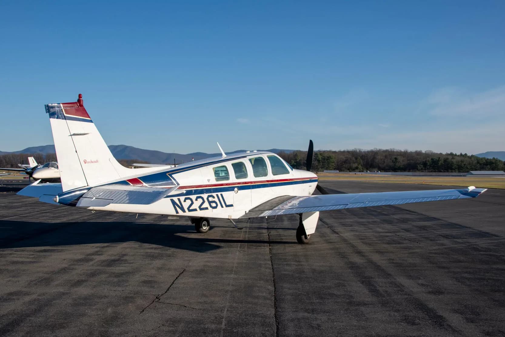 A white Beechcraft Bonanza N2261L with blue and red stripes parked on an airfield, viewed from the rear-left.