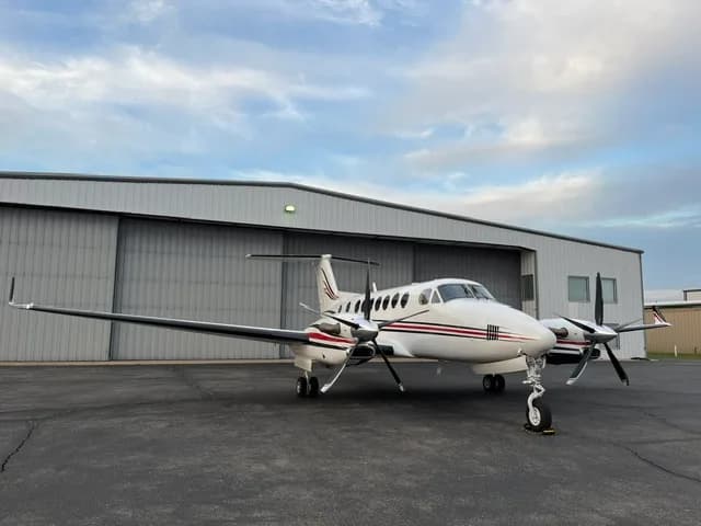 A white Beechcraft King Air with red and black stripes parked on an tarmac in front of a hangar, viewed from the front-right.