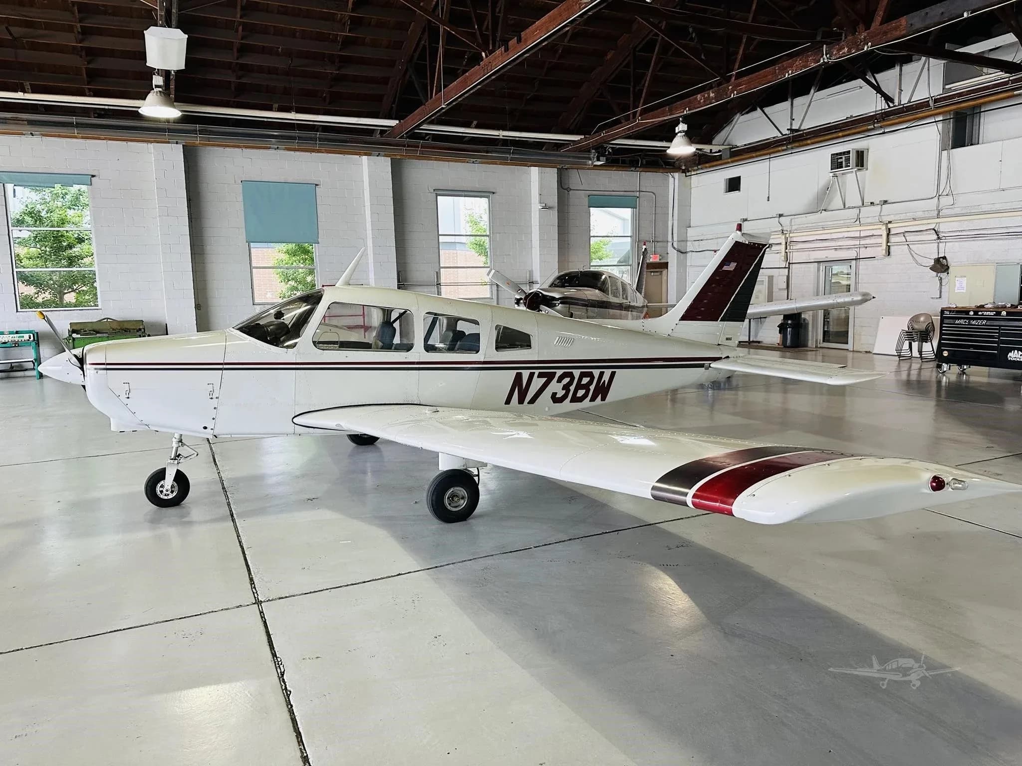 A white single-engine aircraft with maroon and black stripes, registration N73BW, viewed from the left side in a hangar.