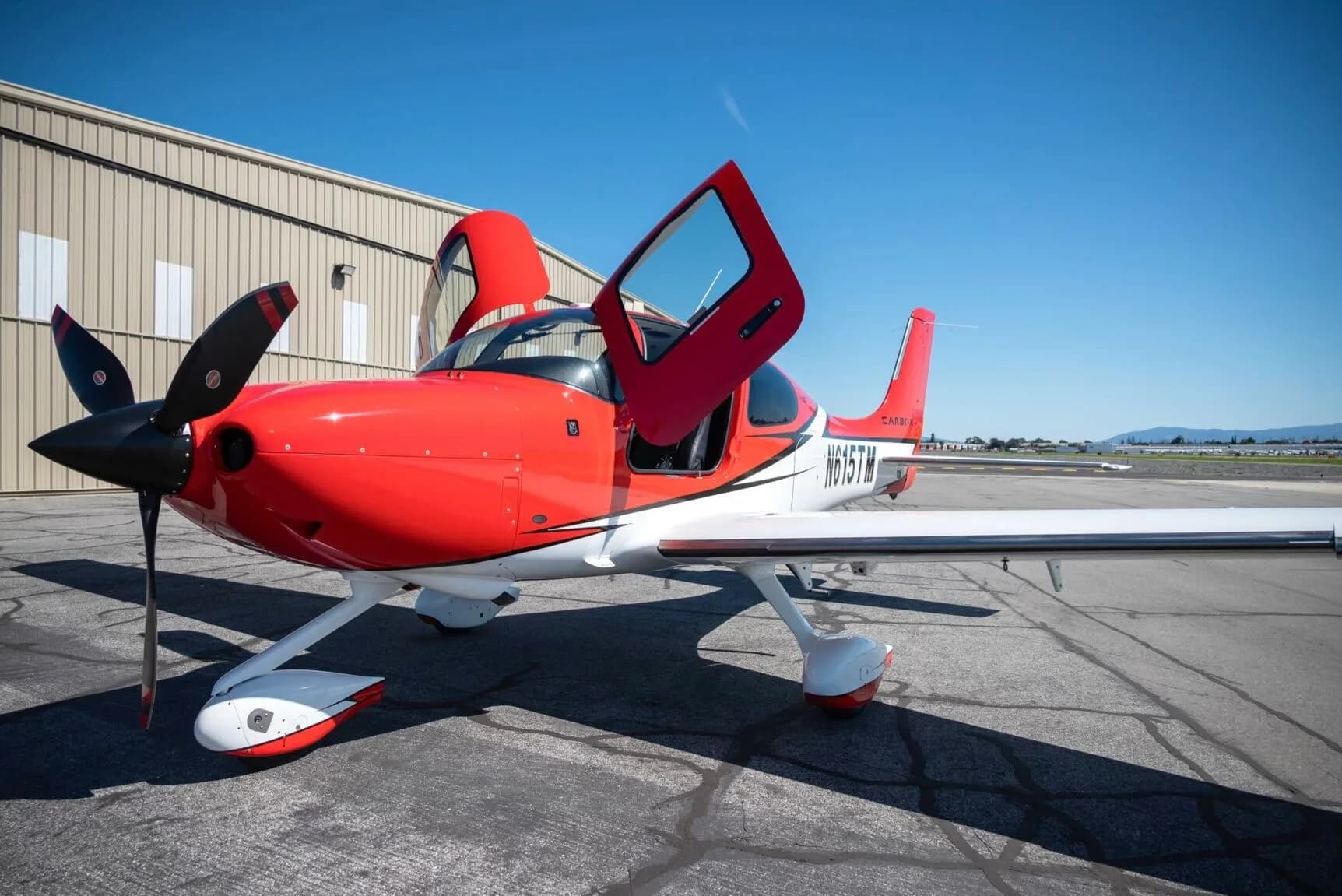 Low-angle front-side view of a red and white N615TM aircraft with its doors open next to a hangar.